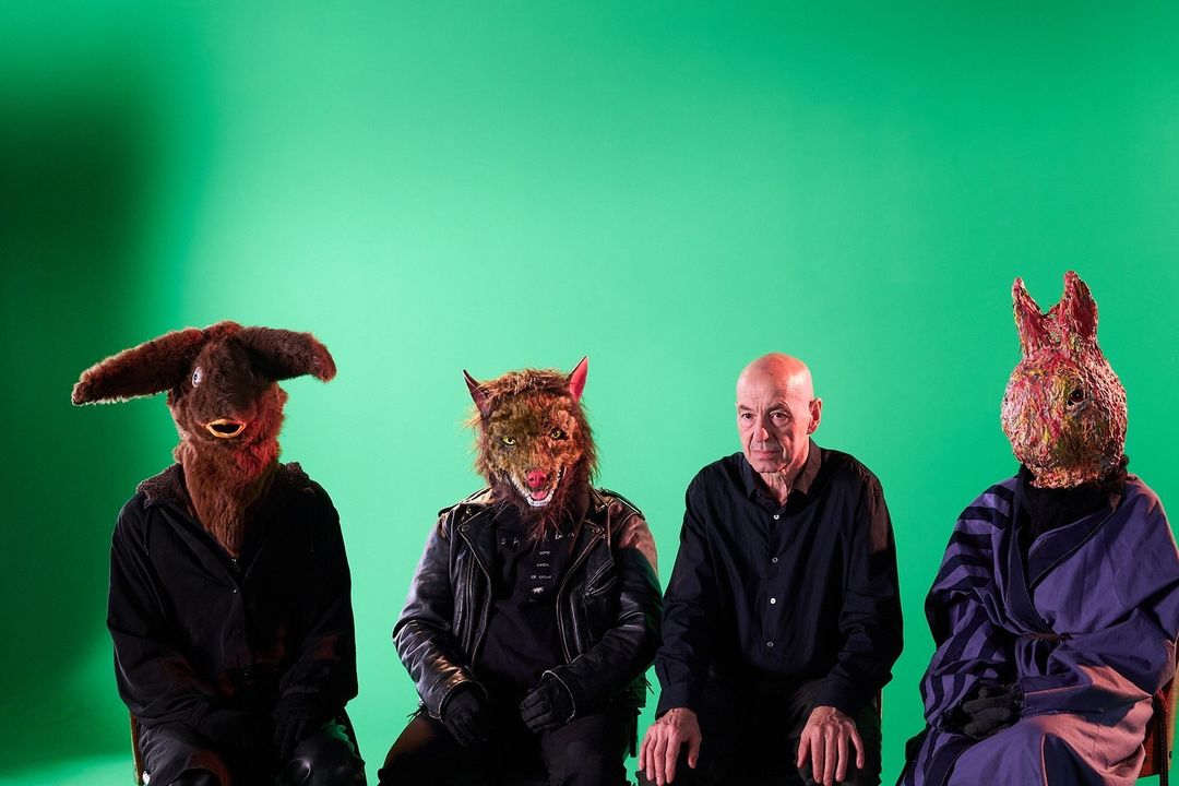 Four figures seated against a green backdrop: one human, and three wearing animal masks: a bull, wolf, and rabbit.
