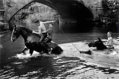 Black and white photograph of two young boys riding a horse through a river beneath a stone bridge