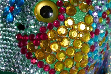 Close-up macro shot of a sculpture encrusted with colorful sparkling rhinestones, featuring a prominent yellow and black eye.