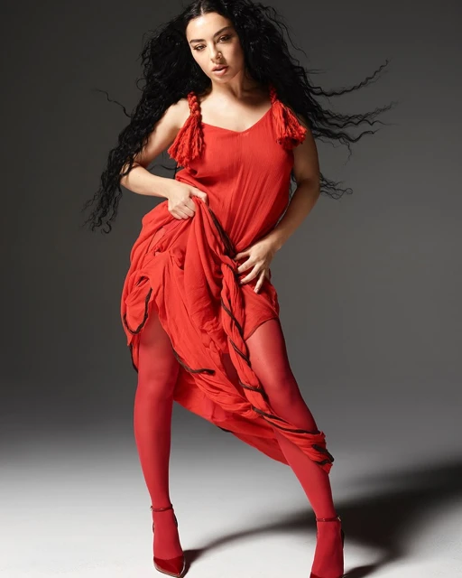Woman with long dark hair in a red dress and matching stockings and shoes posing against a gray background in a studio setting.