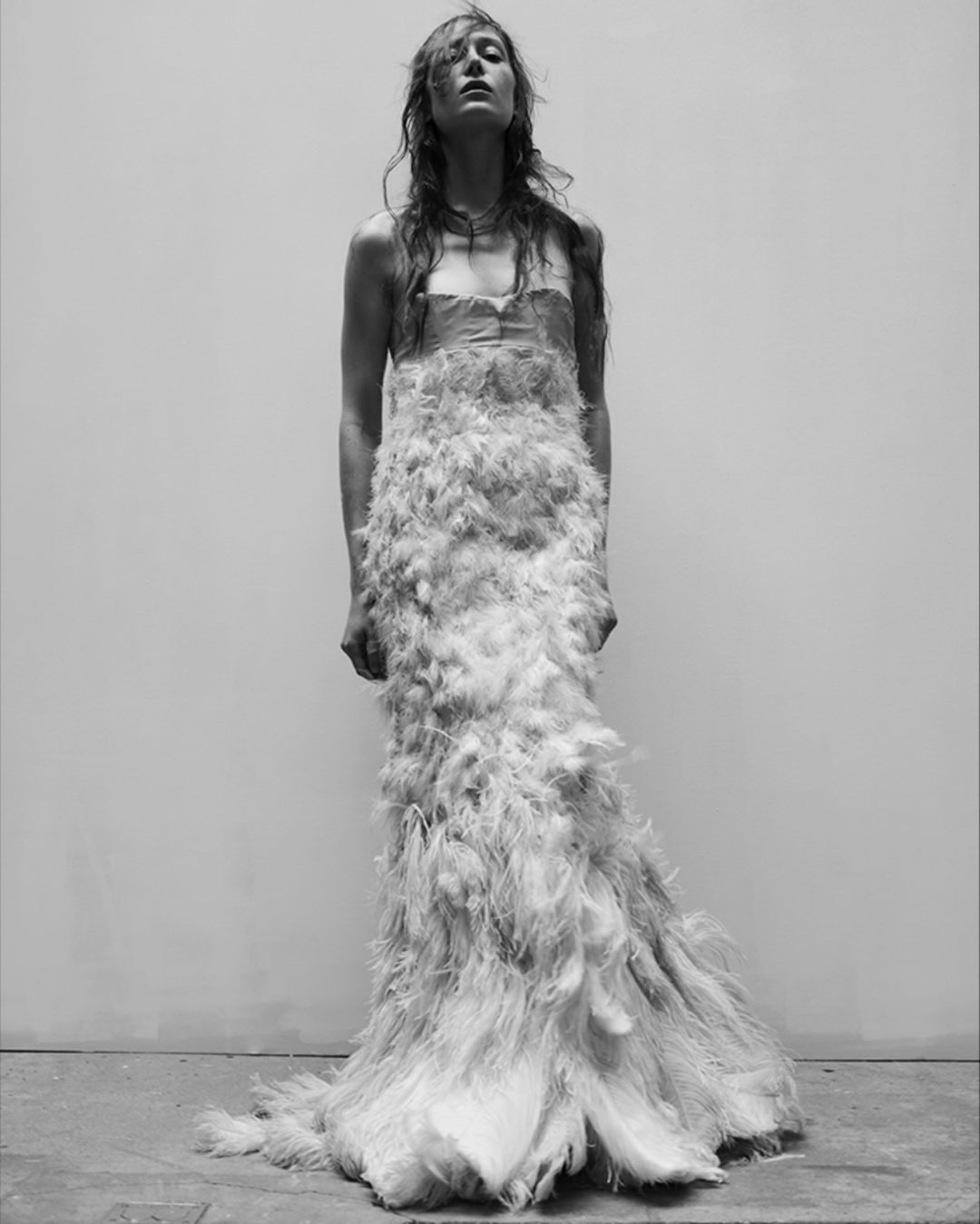 A slender woman with long hair is wearing a feathered gown in this striking black and white studio fashion shot, capturing elegance.