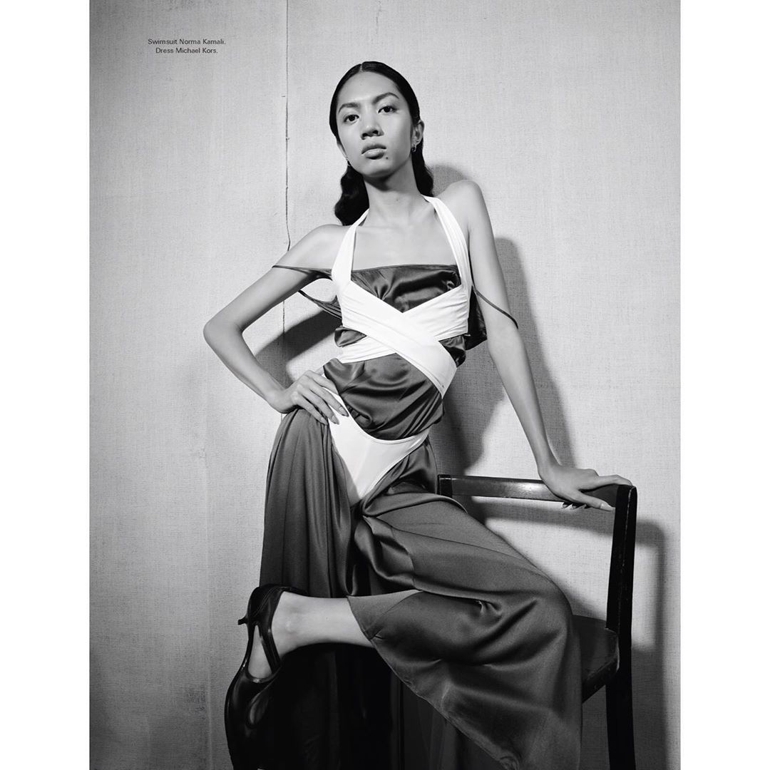 Fashionable woman strikes pose in sleek dress, resting hand on black chair. Black and white medium shot featuring dramatic lighting.
