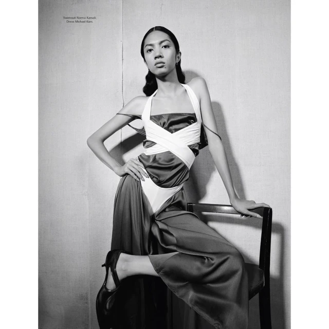 Fashionable woman strikes pose in sleek dress, resting hand on black chair. Black and white medium shot featuring dramatic lighting.