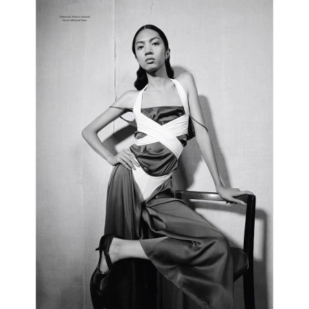 Fashionable woman strikes pose in sleek dress, resting hand on black chair. Black and white medium shot featuring dramatic lighting.