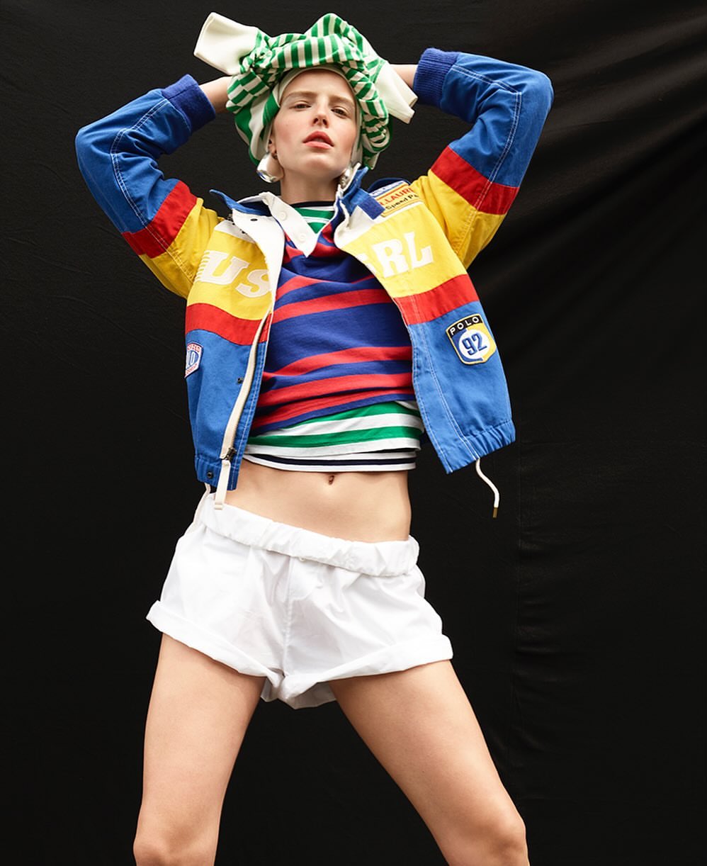 Fashion editorial image featuring a female model in a blue, yellow, and red racing jacket, white shorts, and striped top against a black backdrop.
