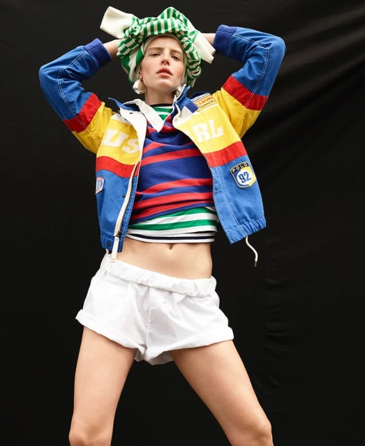 Fashion editorial image featuring a female model in a blue, yellow, and red racing jacket, white shorts, and striped top against a black backdrop.