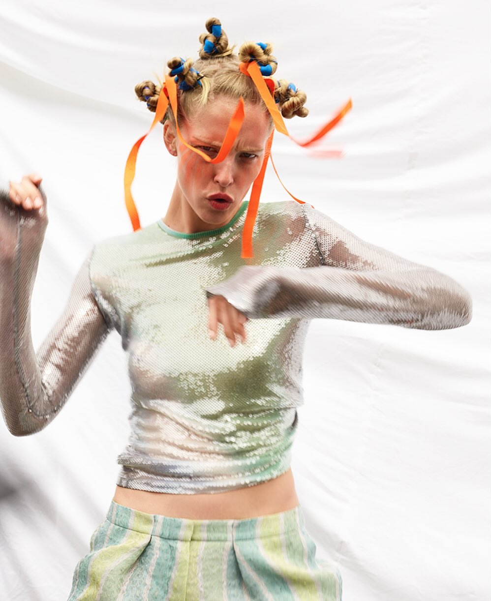 Woman in sparkly top and striped pants poses against a white backdrop. The model has an interesting hairstyle and orange makeup around her eyes.