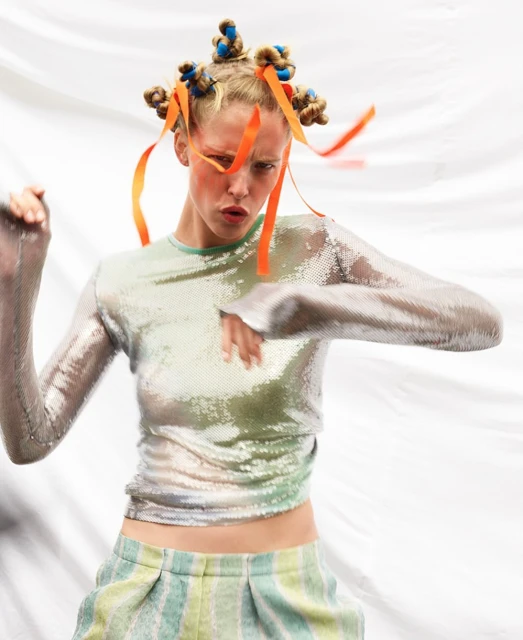 Woman in sparkly top and striped pants poses against a white backdrop. The model has an interesting hairstyle and orange makeup around her eyes.