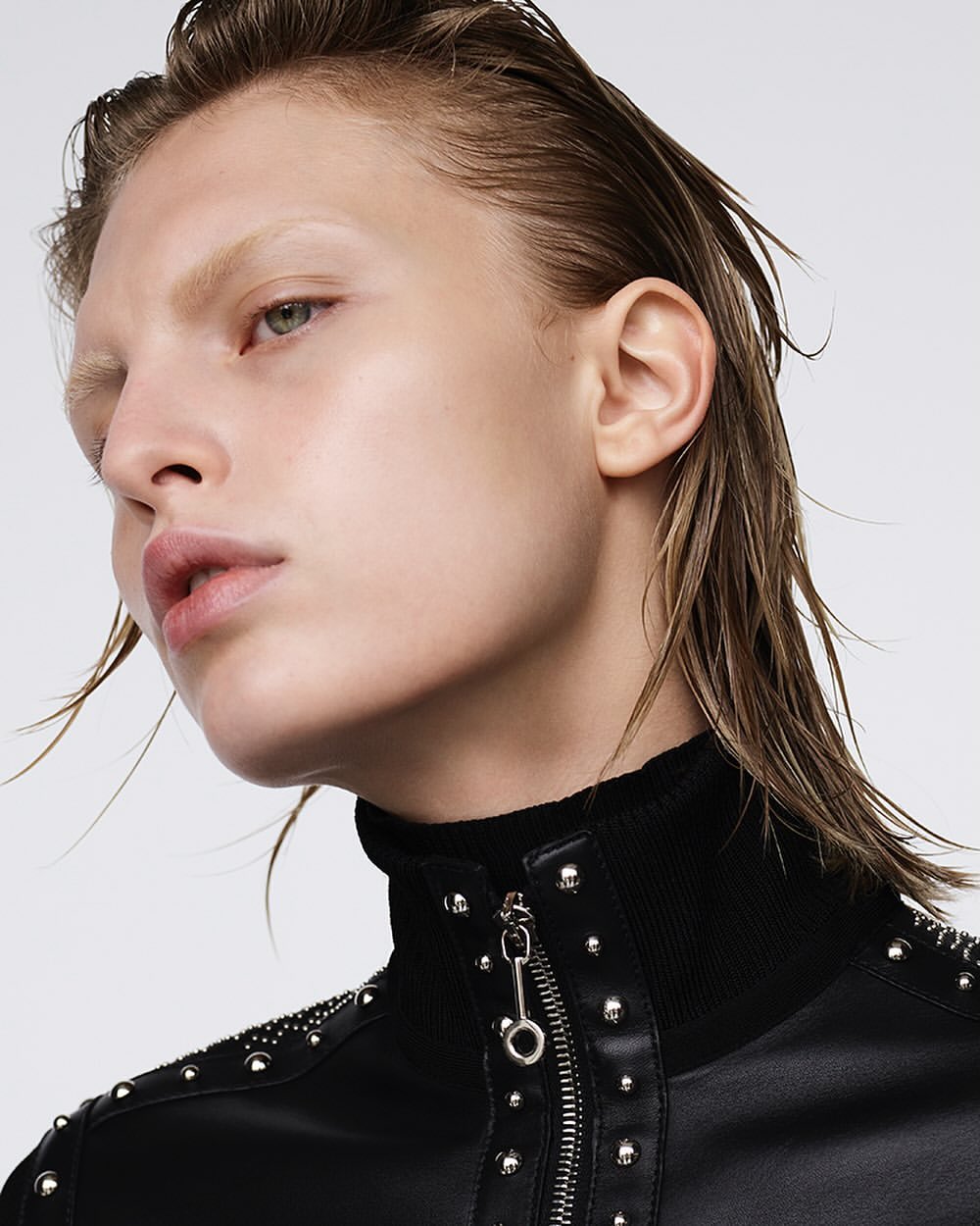 Close-up studio shot of a fair-skinned adult woman with wet blonde hair, wearing a black jacket with silver studs and zipper details, against a white background.