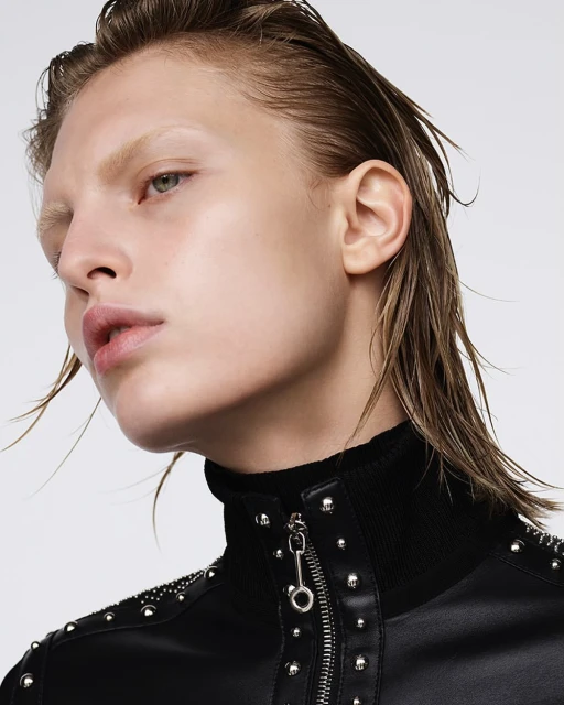 Close-up studio shot of a fair-skinned adult woman with wet blonde hair, wearing a black jacket with silver studs and zipper details, against a white background.