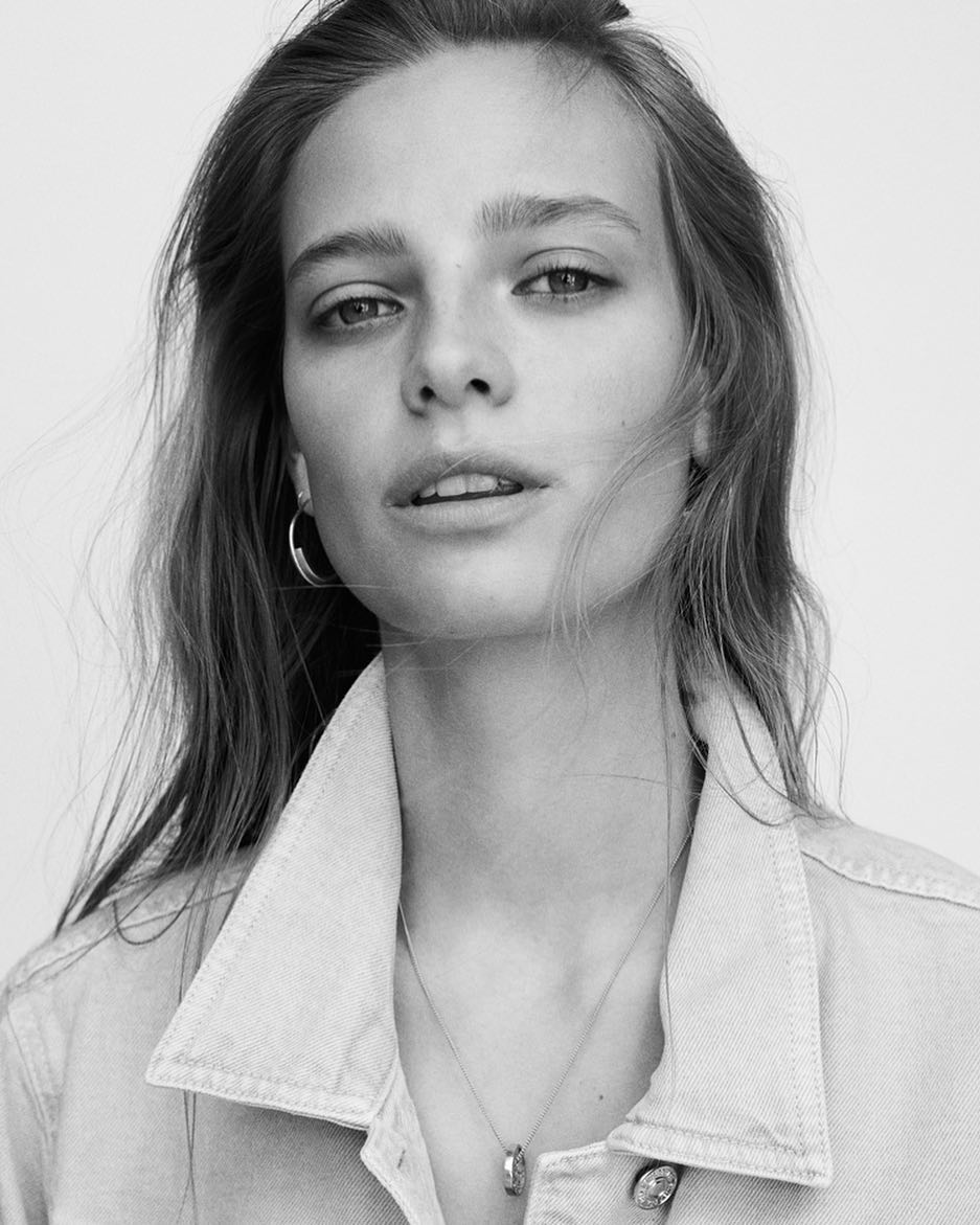 Black and white portrait of a young adult female with long brown hair wearing a denim jacket and silver necklace in a studio setting.