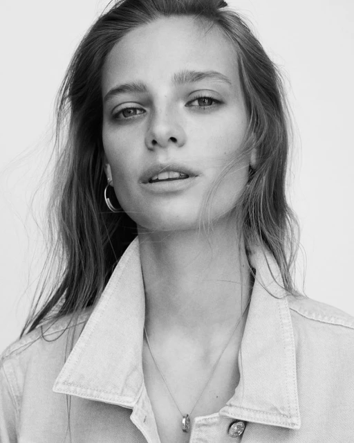 Black and white portrait of a young adult female with long brown hair wearing a denim jacket and silver necklace in a studio setting.