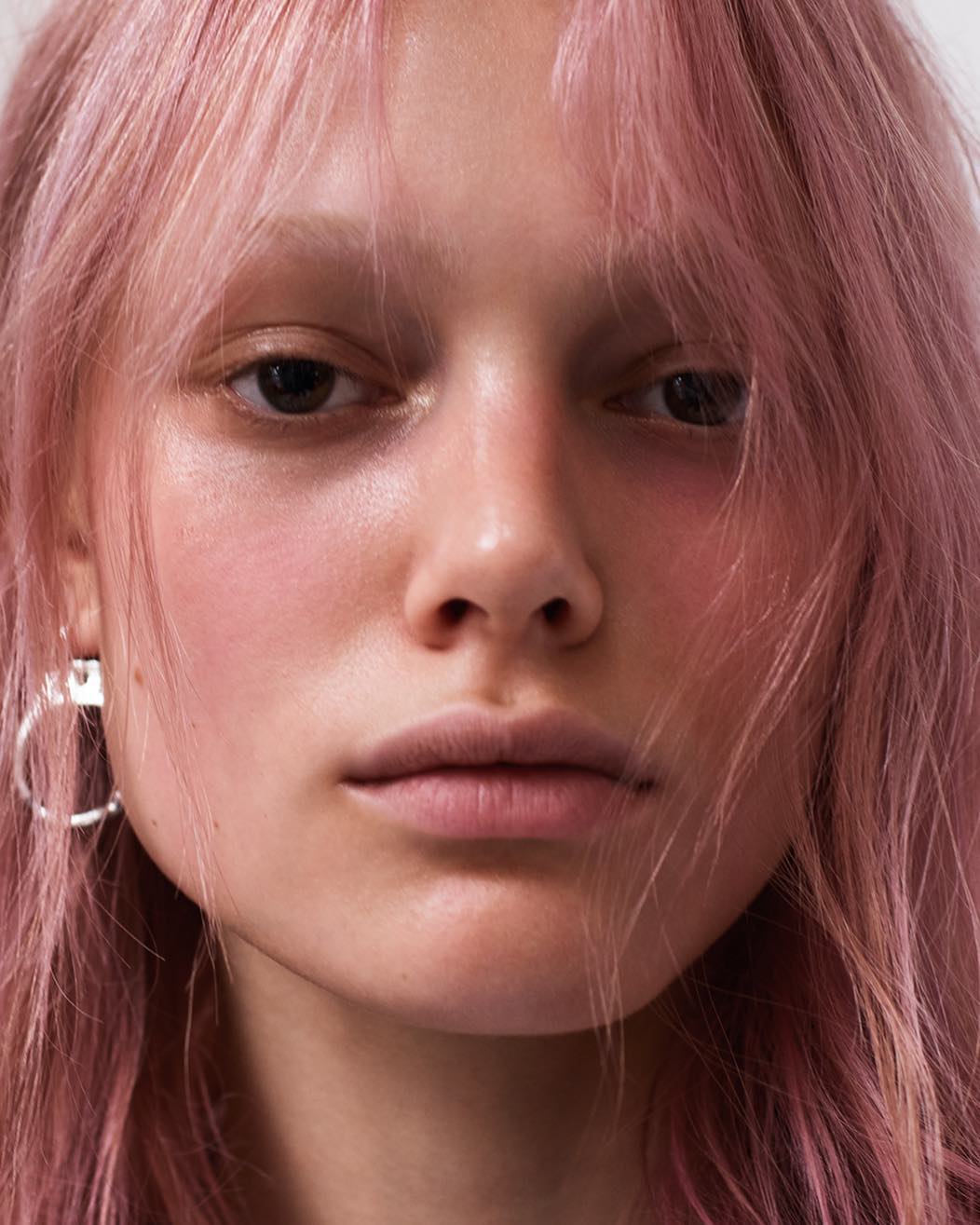 Close-up portrait of an adult female with pink hair and fair skin, wearing silver earrings, set against a soft, neutral background.