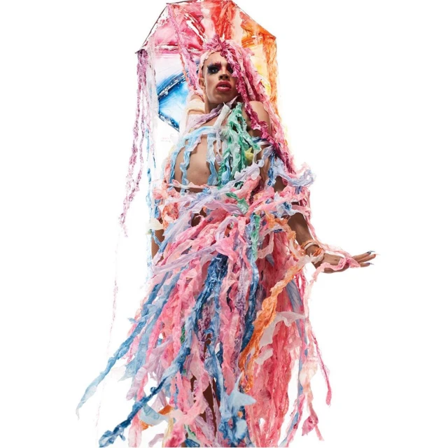 A person in drag wearing colorful plastic material and makeup, standing in a brightly lit indoor studio for a fashion-forward portrait.