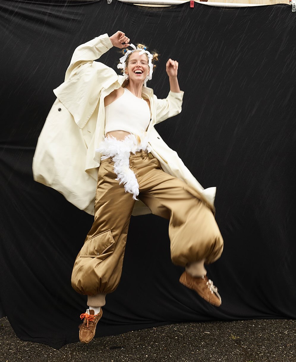 A cheerful woman in an off-white top and cream overcoat jumps indoors, wearing brown pants, and shoes against a black backdrop.