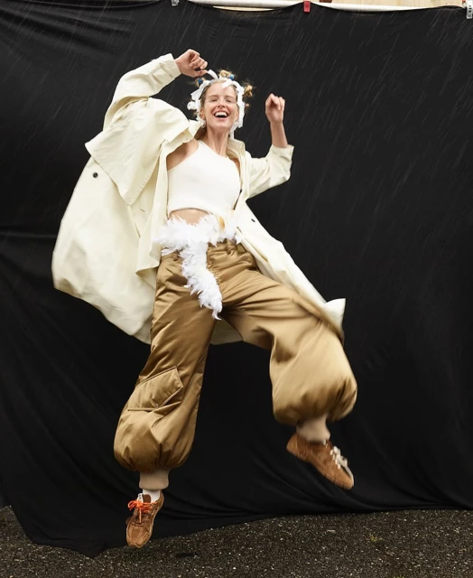 A cheerful woman in an off-white top and cream overcoat jumps indoors, wearing brown pants, and shoes against a black backdrop.