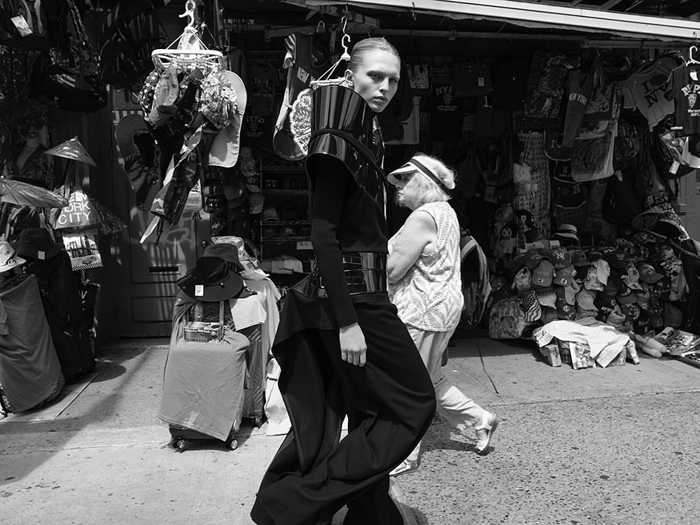 Fashionable adult female posing in a high-fashion outfit on a city sidewalk with a secondary adult female in the background.