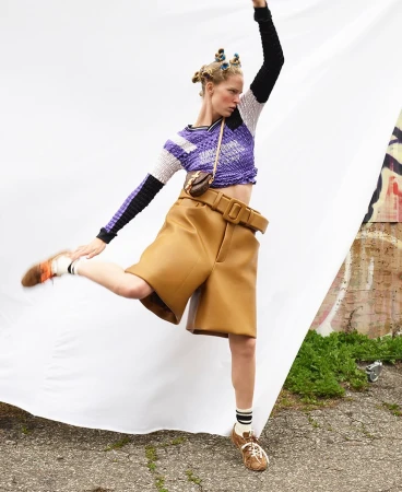 Model with styled hair in purple textured top and tan shorts posing against a white backdrop.