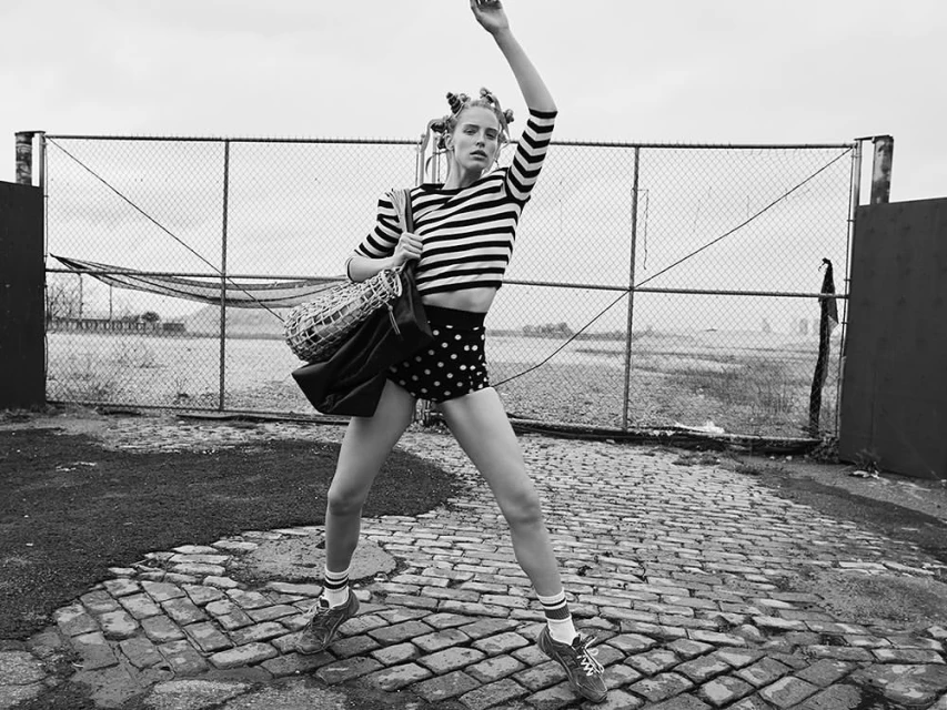 A woman in vintage fashion stands with arms raised on a cobblestone street, showcasing a black and white striped top and polka dot shorts.