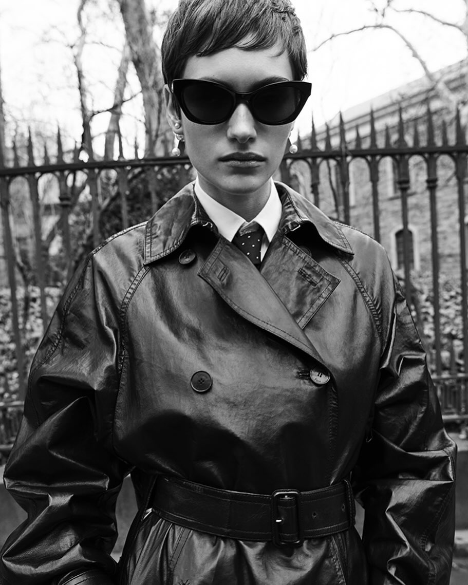 Monochrome portrait of a woman in a trench coat and sunglasses standing outdoors against a wrought iron fence and building.