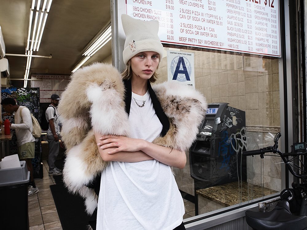 A young woman in a knitted hat and faux fur stole poses in front of a New York City pizza restaurant window with a menu board.