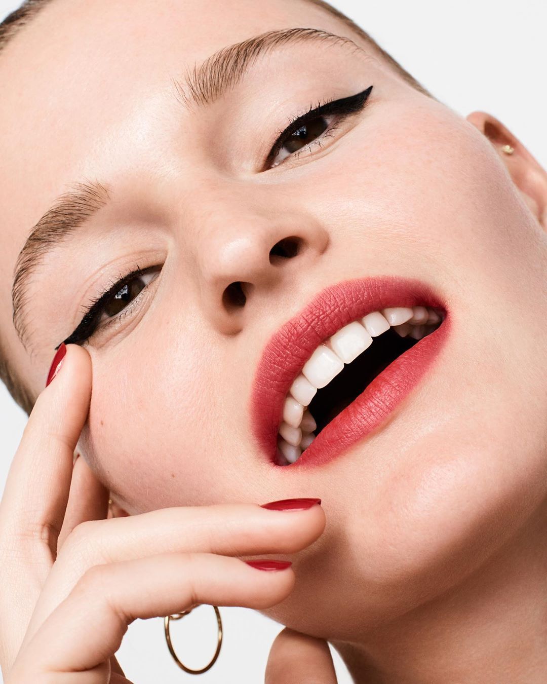 Close-up portrait of a fair-skinned woman wearing makeup with cat eyeliner and red lipstick, posing against a neutral background.