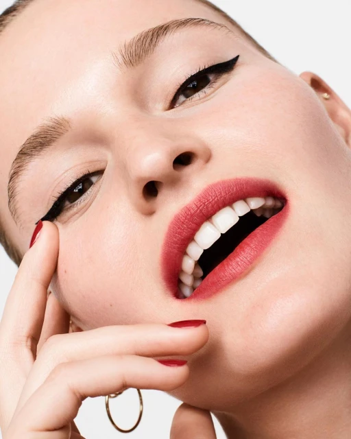 Close-up portrait of a fair-skinned woman wearing makeup with cat eyeliner and red lipstick, posing against a neutral background.