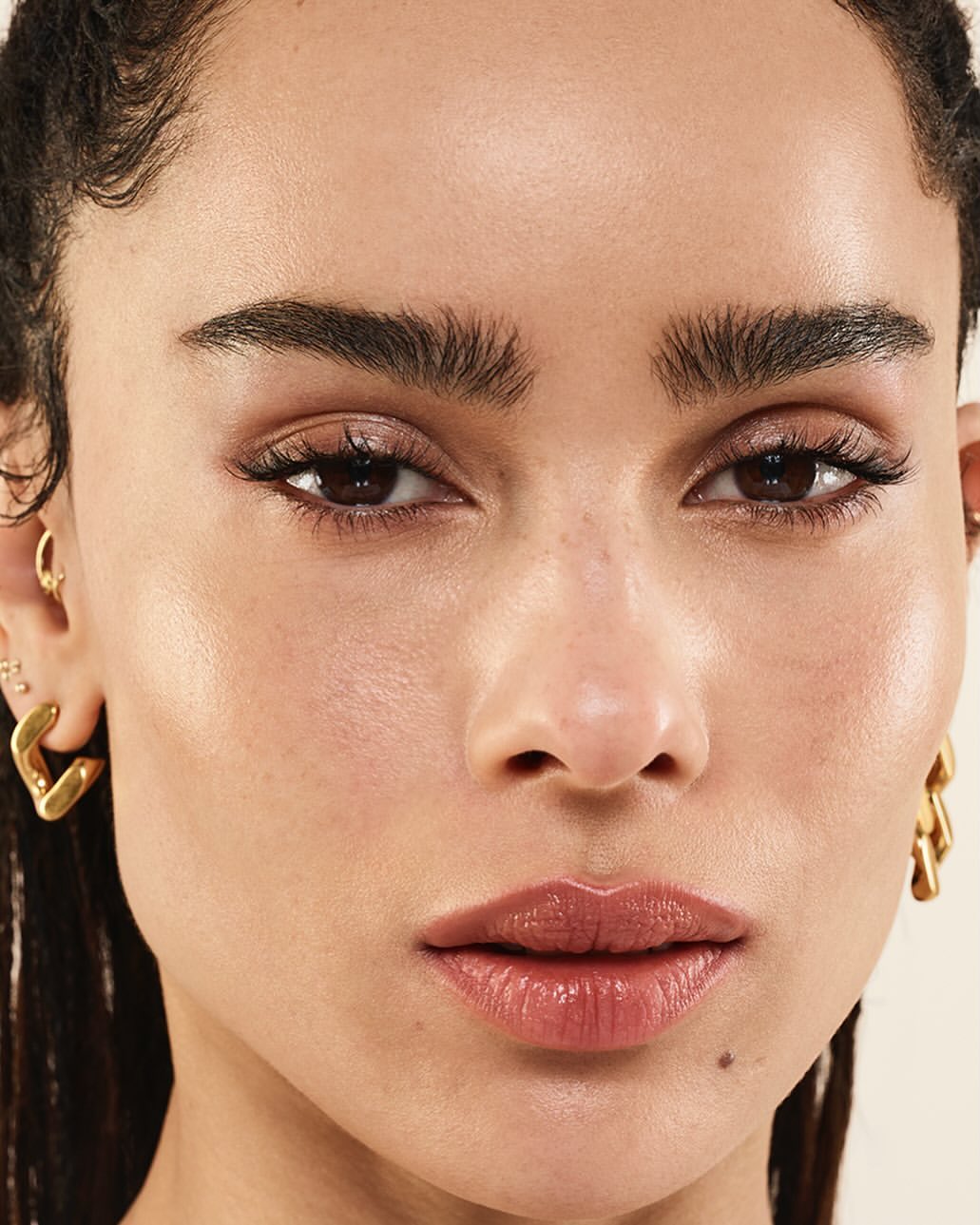 Close-up portrait of a brunette adult female with gold earrings, neutral makeup, and defined eyebrows against a neutral background.