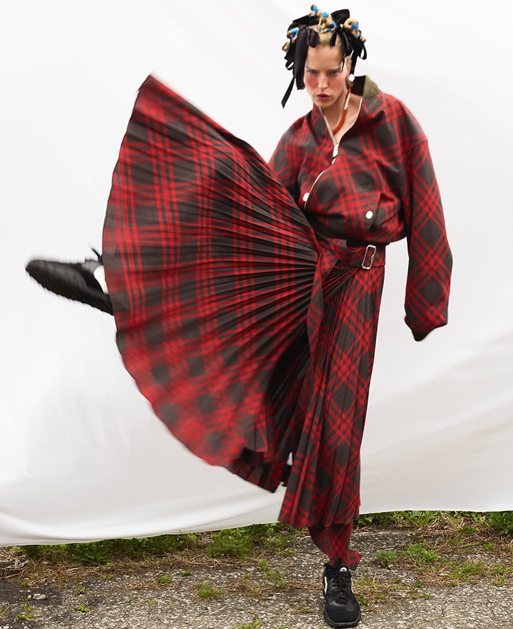 Adult female poses in a red and black plaid outfit, one leg raised, against a white fabric background and grassy ground outdoors.