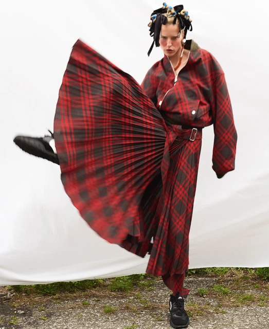 Adult female poses in a red and black plaid outfit, one leg raised, against a white fabric background and grassy ground outdoors.