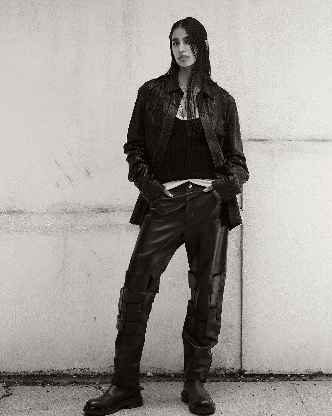 Monochrome fashion shot: a woman poses in a leather jacket, tank top, and unique pants with straps, standing against a textured concrete wall.