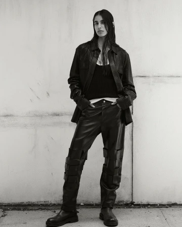 Monochrome fashion shot: a woman poses in a leather jacket, tank top, and unique pants with straps, standing against a textured concrete wall.