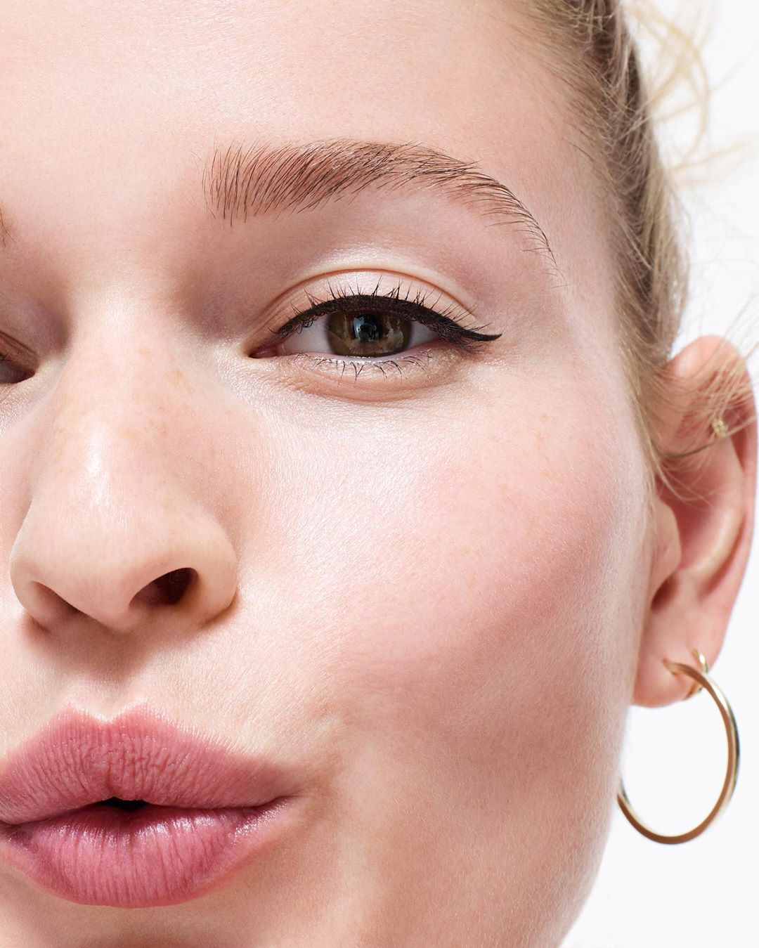 Close-up of a young adult woman's face with light skin, dark eyeliner, and pink lips, set against a bright, neutral background.