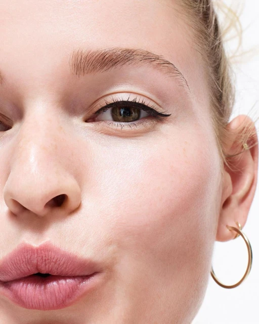 Close-up of a young adult woman's face with light skin, dark eyeliner, and pink lips, set against a bright, neutral background.