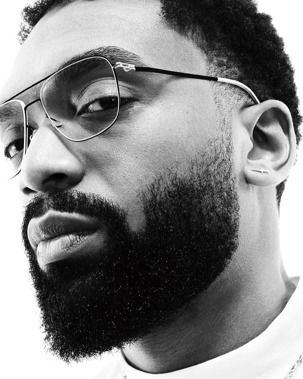 Black and white portrait of a stylish adult male with a beard and eyeglasses against a bright white backdrop in a photo studio.