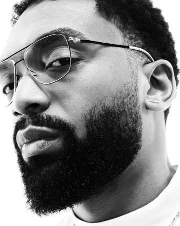 Close-up portrait of a Black man with a full beard wearing wire-frame glasses and a white shirt against a seamless background.