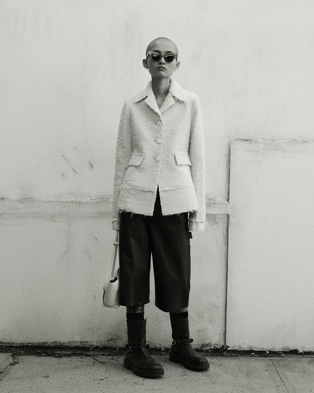 A bald woman poses in a tweed jacket, dark shorts, and boots, carrying a cream purse against a mottled white wall outdoors.