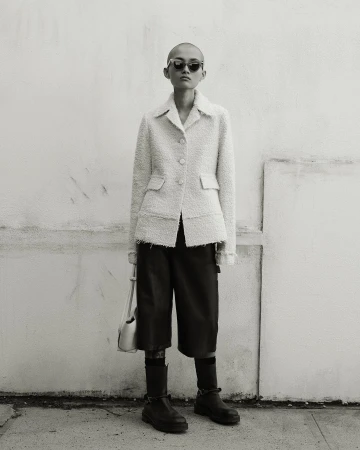 Model with shaved head wearing dark sunglasses, textured light jacket, dark bermuda shorts, and boots.