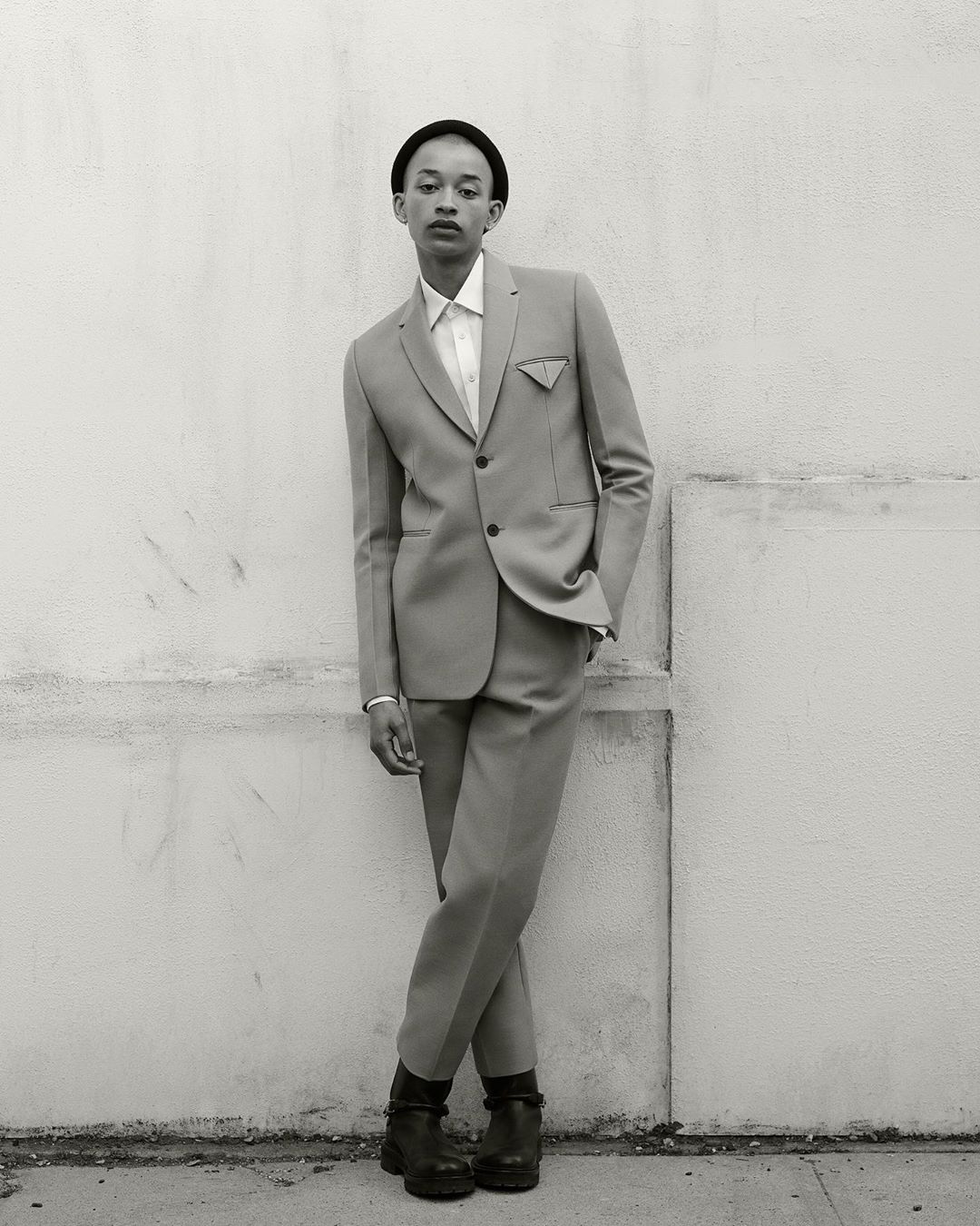 Monochrome portrait of a slender person posing in a light grey suit with a black beanie against a weathered white wall outdoors.