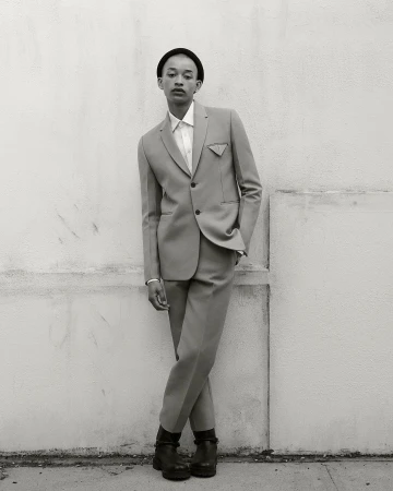 Monochrome portrait of a slender person posing in a light grey suit with a black beanie against a weathered white wall outdoors.