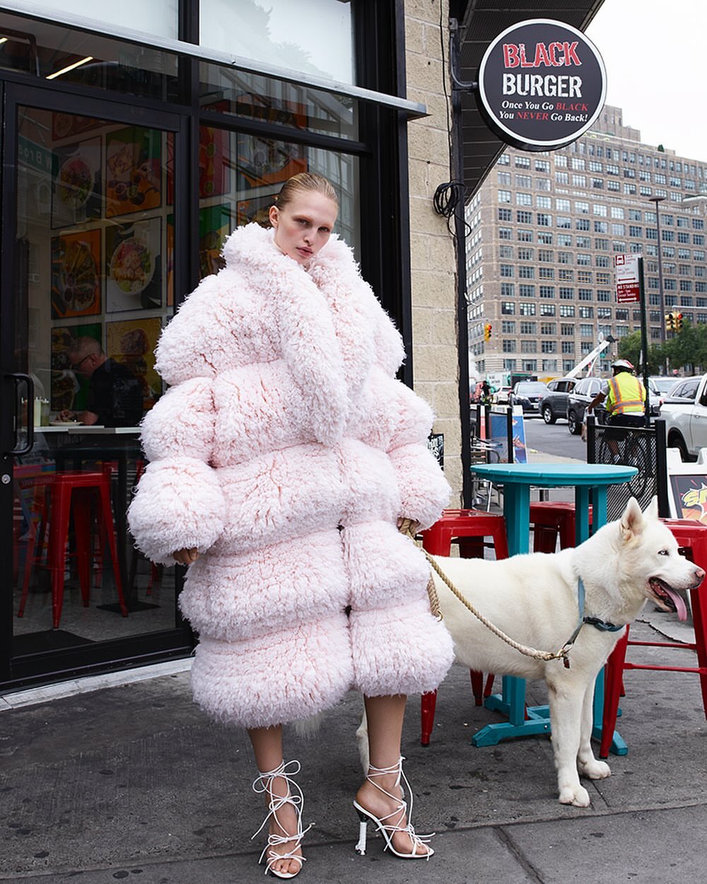Model in oversized textured pink coat standing on a city sidewalk next to a white dog wearing a leash by Amy Troost