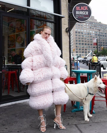 Model in oversized textured pink coat standing on a city sidewalk next to a white dog wearing a leash by Amy Troost