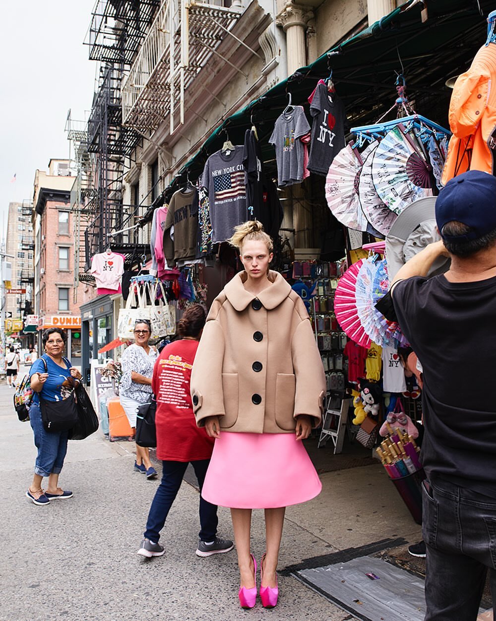 Fashion shot of a model in a tan coat and pink skirt standing on a city sidewalk with souvenir shops and people in the background.