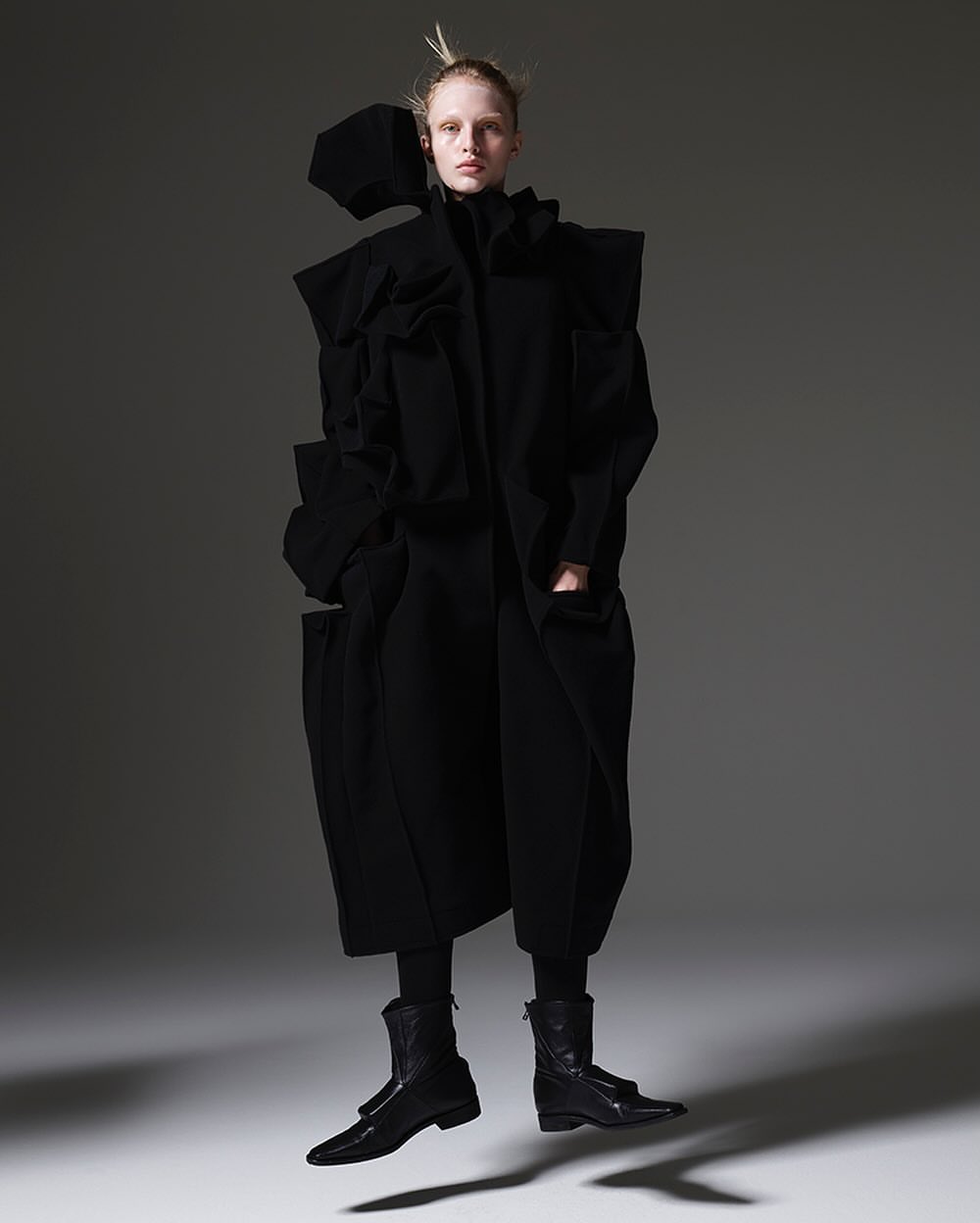 A fashion photograph showcasing an adult female model in an avant-garde black outfit, captured in a full shot against a grey backdrop.