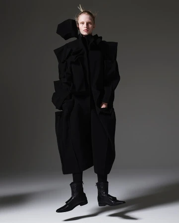 A fashion photograph showcasing an adult female model in an avant-garde black outfit, captured in a full shot against a grey backdrop.