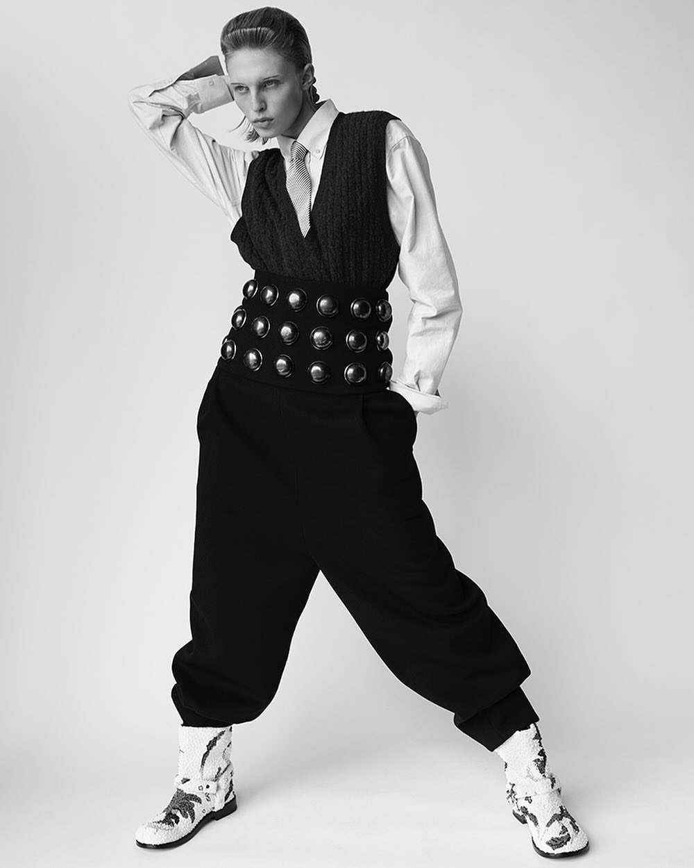 Monochrome fashion shot: a model poses in a white shirt, knitted vest, wide belt, oversized trousers, and patterned boots against a plain background.
