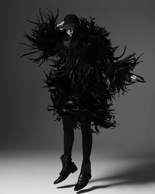 Black and white fashion shot of a woman in a feather coat, hat, leggings, and boots jumping in a gray photo studio.