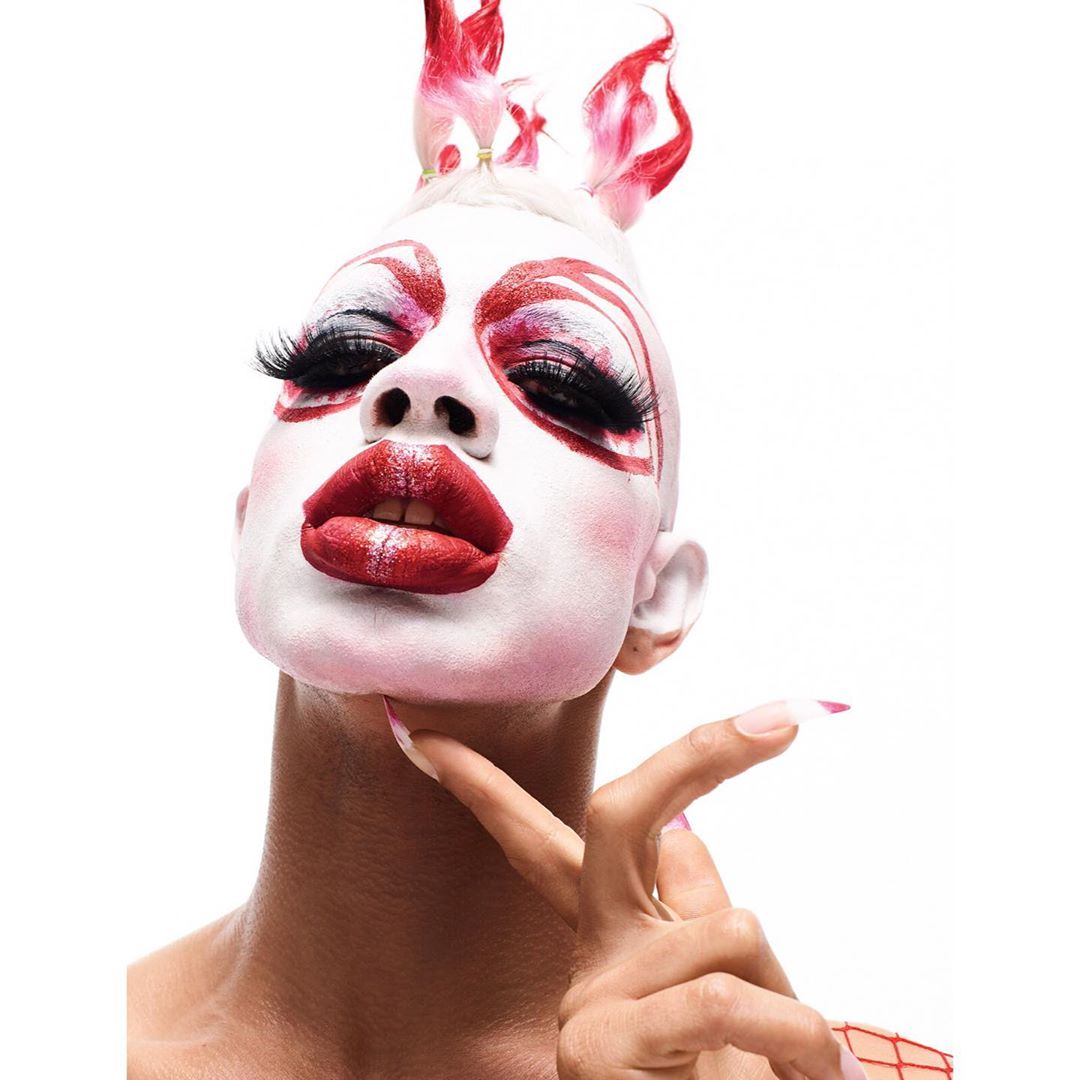 Portrait of a person with white face paint, red accents, exaggerated eyelashes, and artistic red and white headpiece, facing the camera.