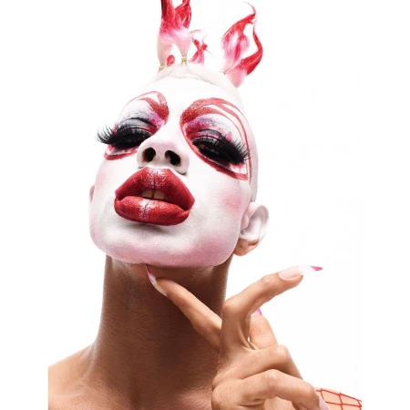 Portrait of a person with white face paint, red accents, exaggerated eyelashes, and artistic red and white headpiece, facing the camera.
