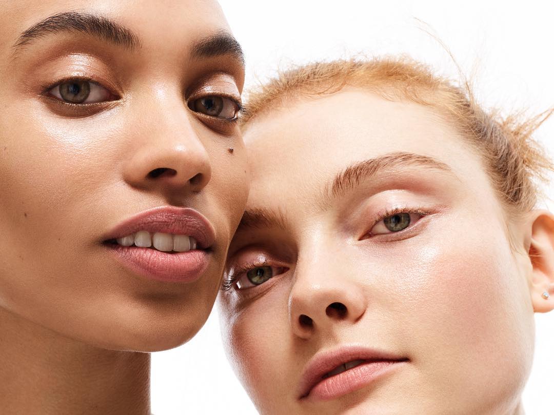 Close-up studio portrait of two women with fair skin tones, soft lighting, and subtle makeup against a stark white backdrop.